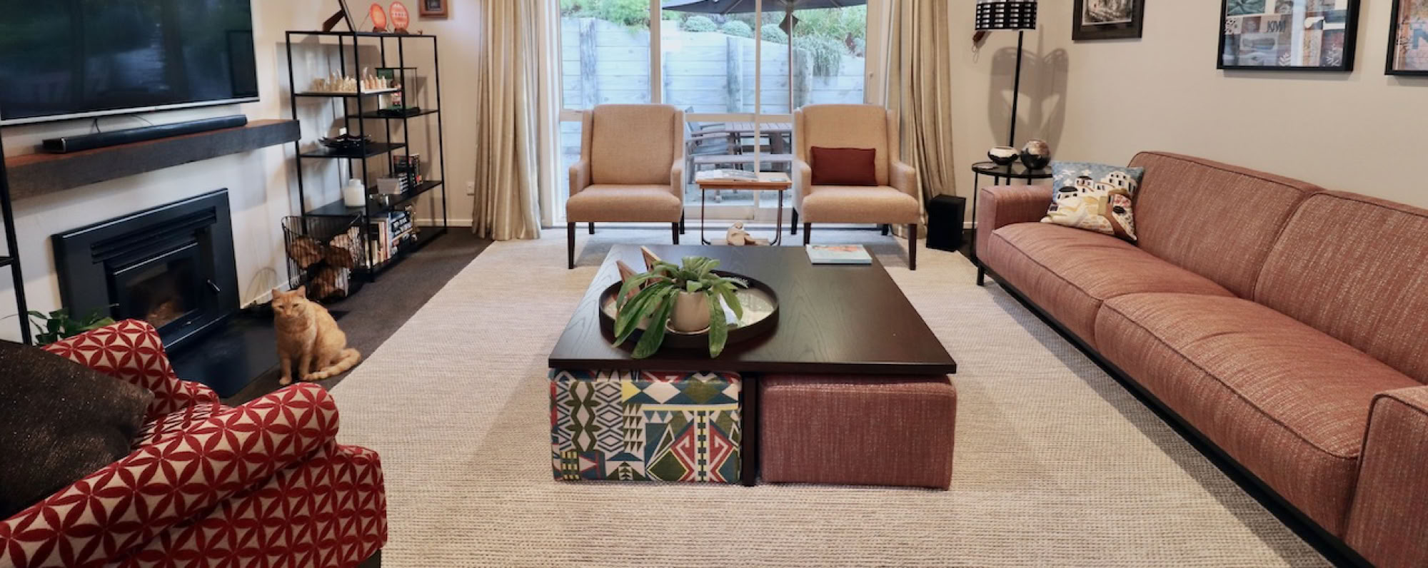 Studio 28 Interio Design Wellington Zealand. Creating wow interiors that are easy on the eye. Living room with light coloured rug over dark carpet. sofa and 3 chairs with coffee table and ottomans. TV wall is balalnced with shelving either side.